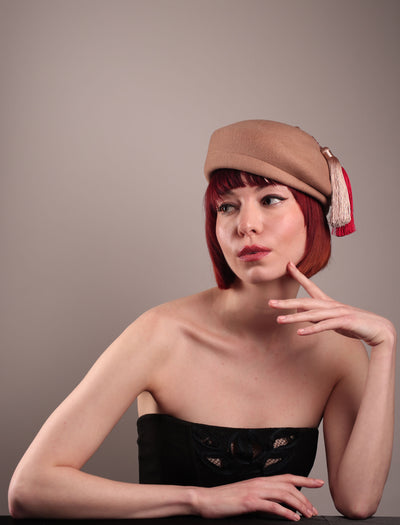 The model is showcasing the rigth front side of the  Elera Felt Beret Hat, made of smooth camel wool felt. This structured beret features a dramatic two-tone tassel in crimson and cream, gently draped to one side from a polished metallic accent. Designed by Merve Bayindir. 