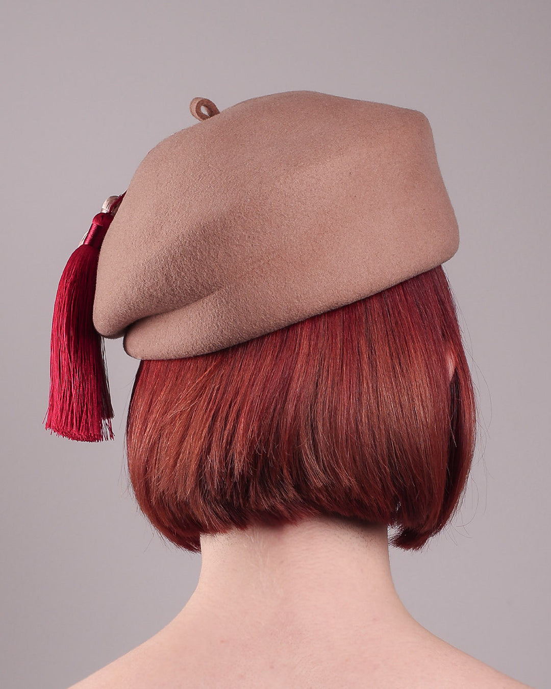 The model is showcasing the back of Elera Felt Beret Hat, made of smooth camel wool felt. This structured beret features a dramatic two-tone tassel in crimson and cream, gently draped to one side from a polished metallic accent. Designed by Merve Bayindir. 