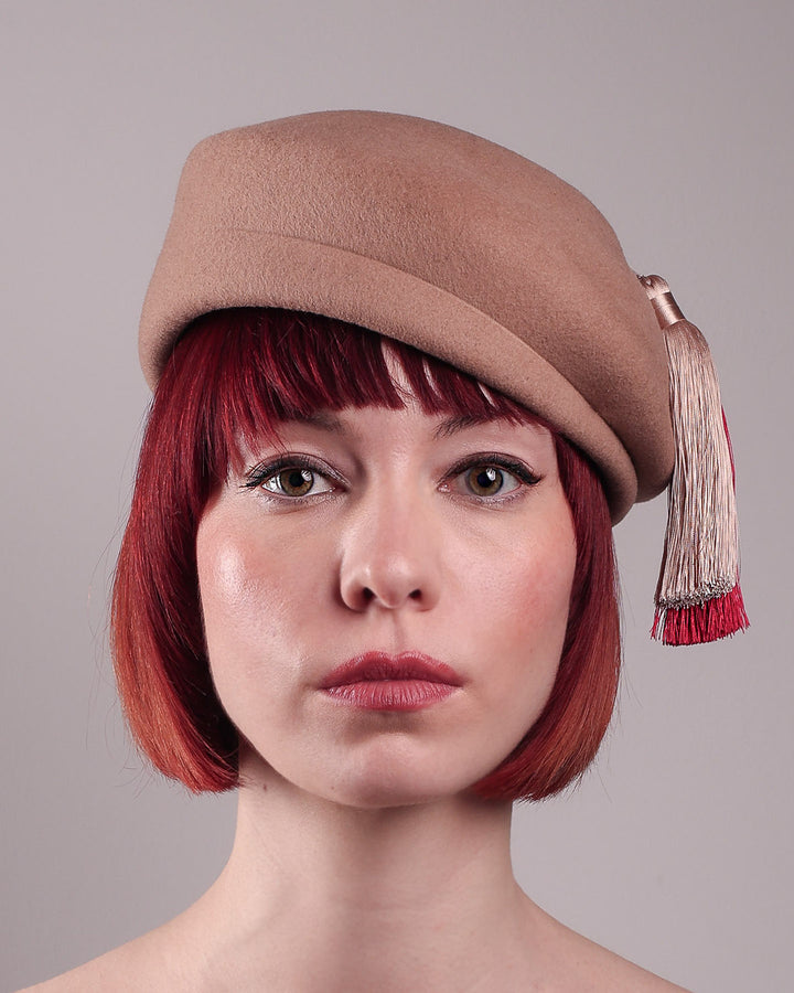 The model is showcasing the front close up of Elera Felt Beret Hat, made of smooth camel wool felt. This structured beret features a dramatic two-tone tassel in crimson and cream, gently draped to one side from a polished metallic accent. Designed by Merve Bayindir. 