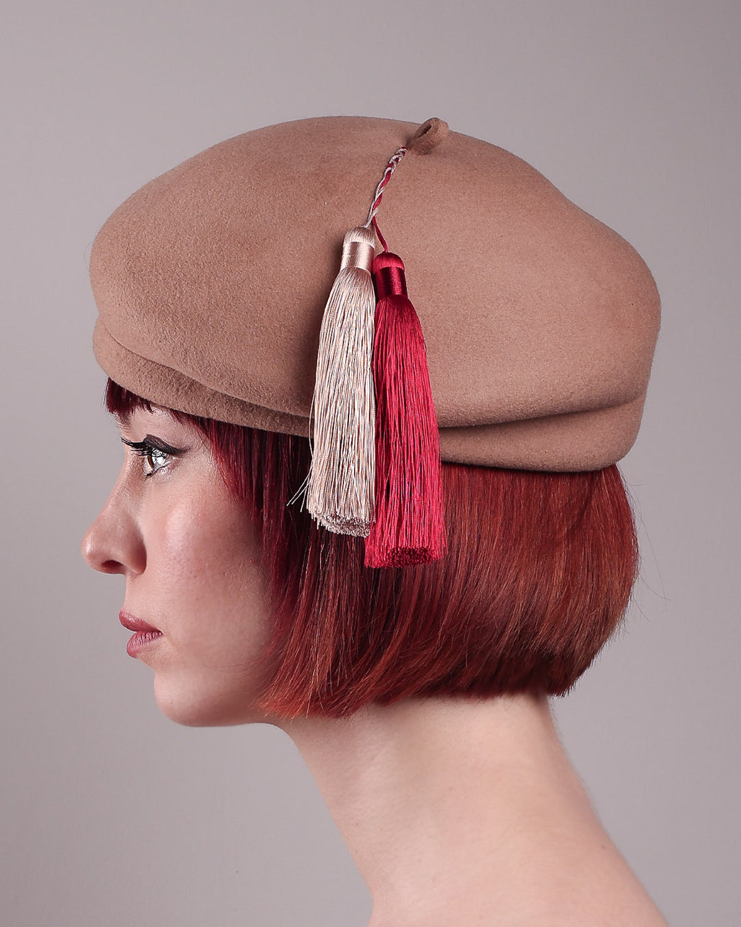 The model is showcasing the right side close up of Elera Felt Beret Hat, made of smooth camel wool felt. This structured beret features a dramatic two-tone tassel in crimson and cream, gently draped to one side from a polished metallic accent. Designed by Merve Bayindir. 