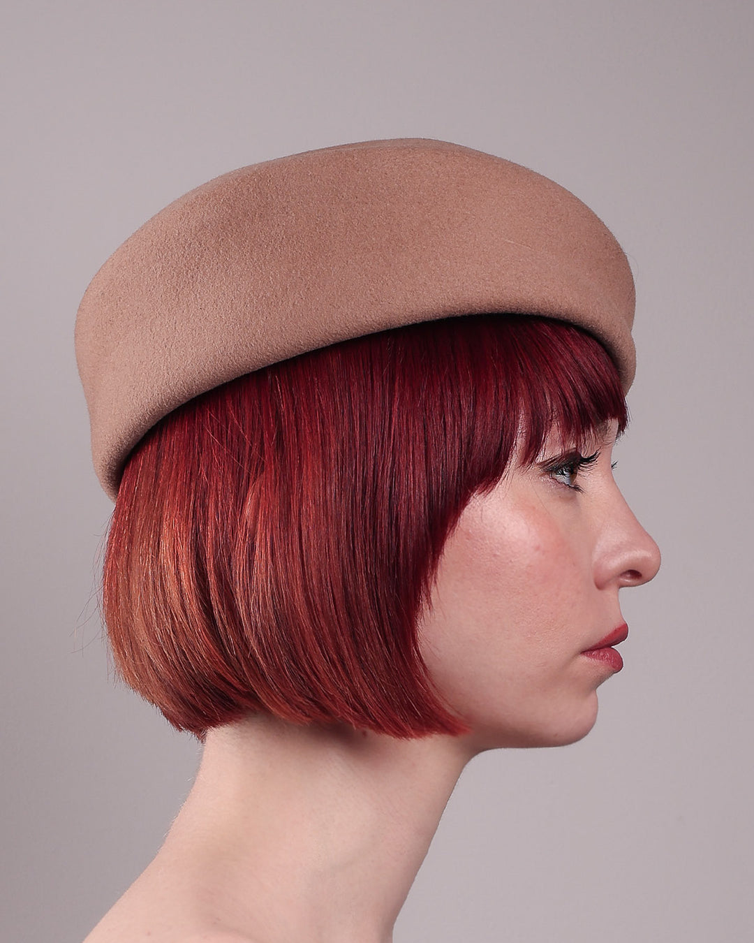 The model is showcasing the right side close up of Elera Felt Beret Hat, made of smooth camel wool felt. This structured beret features a dramatic two-tone tassel in crimson and cream, gently draped to one side from a polished metallic accent. Designed by Merve Bayindir. 