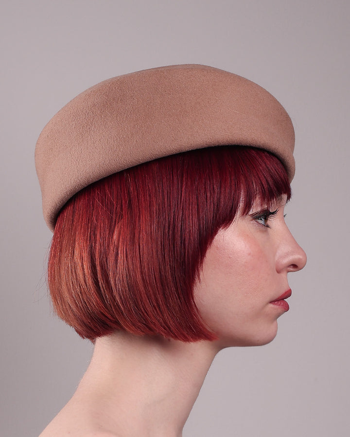 The model is showcasing the right side close up of Elera Felt Beret Hat, made of smooth camel wool felt. This structured beret features a dramatic two-tone tassel in crimson and cream, gently draped to one side from a polished metallic accent. Designed by Merve Bayindir. 