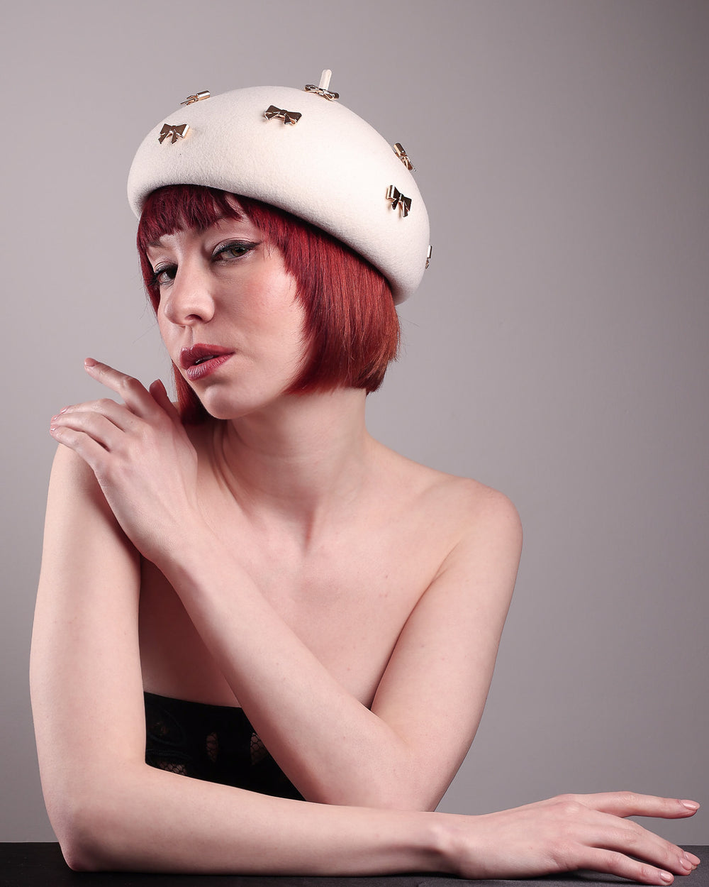 The model poses to showcase the left angle dome-shaped, round Elodie pillbox beret hat, crafted from soft white felt and adorned with gold coloured metal bows. Designed by Merve Bayindir.