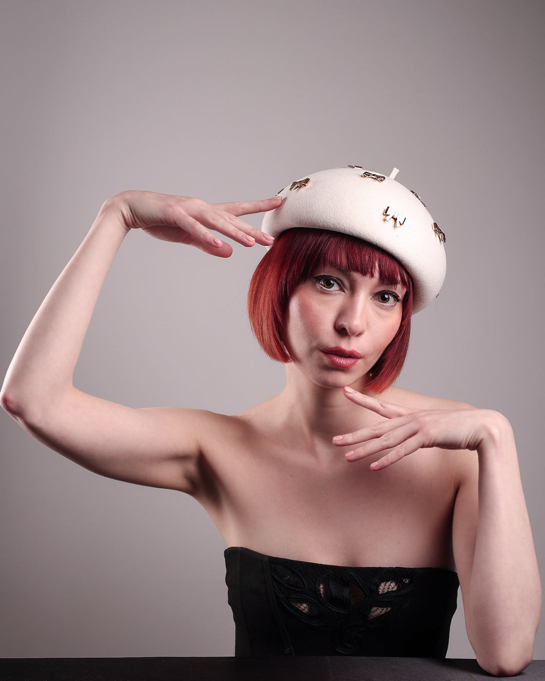 The model poses to showcase the front angle dome-shaped, round Elodie pillbox beret hat, crafted from soft white felt and adorned with gold coloured metal bows. Designed by Merve Bayindir.