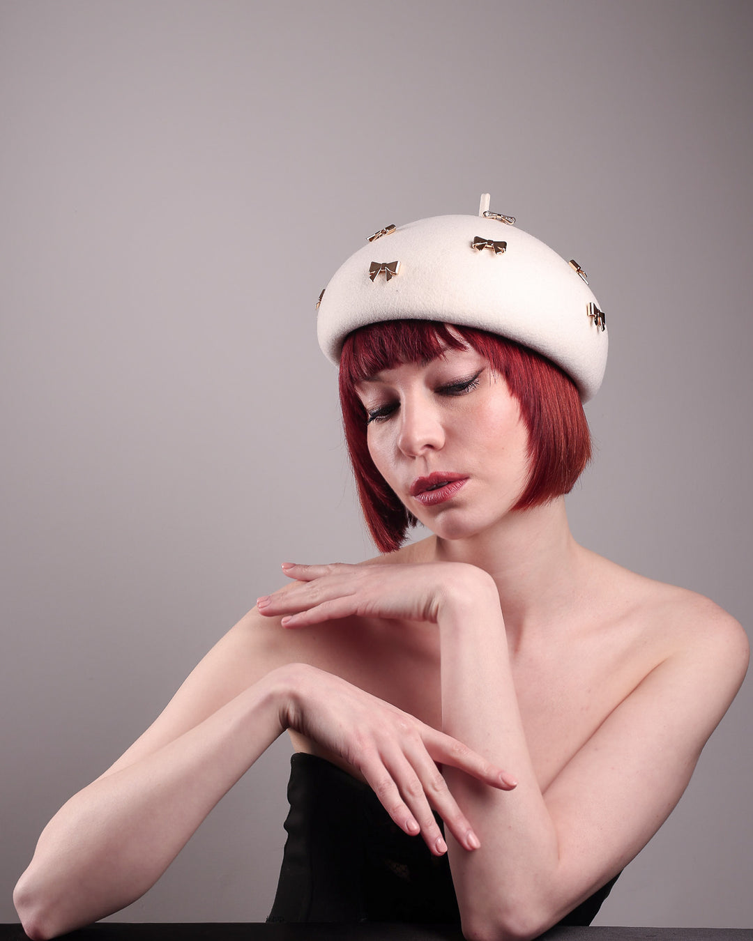 The model poses to showcase the left front angle dome-shaped, round Elodie pillbox beret hat, crafted from soft white felt and adorned with gold coloured metal bows. Designed by Merve Bayindir.