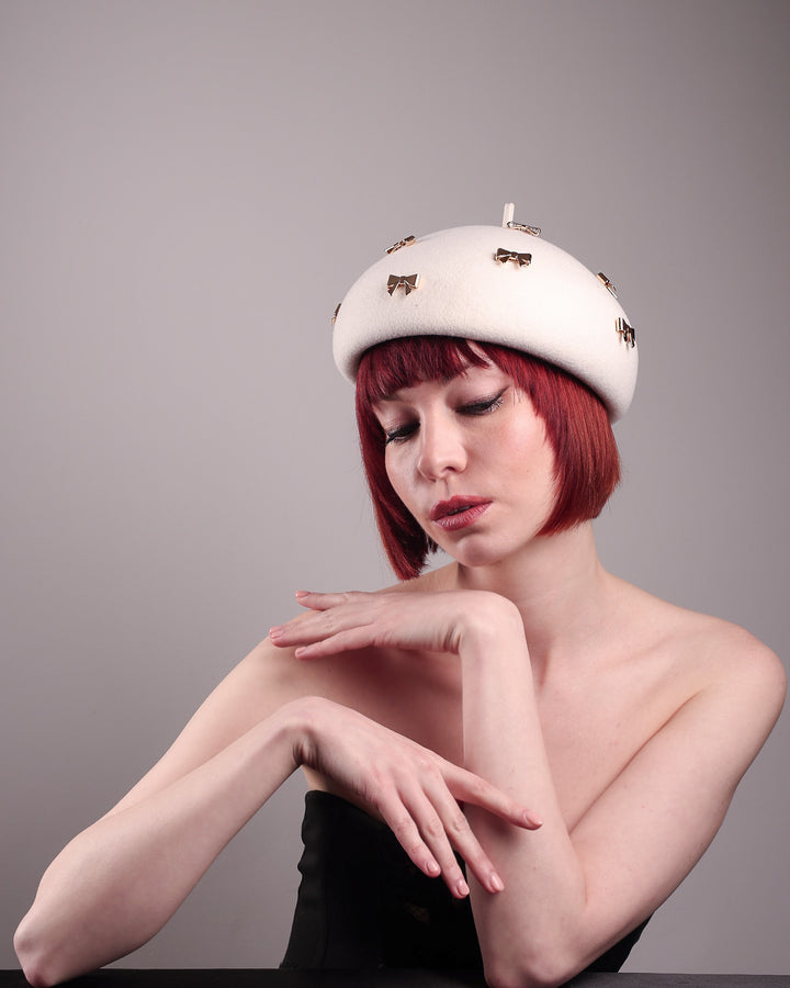 The model poses to showcase the left front angle dome-shaped, round Elodie pillbox beret hat, crafted from soft white felt and adorned with gold coloured metal bows. Designed by Merve Bayindir.