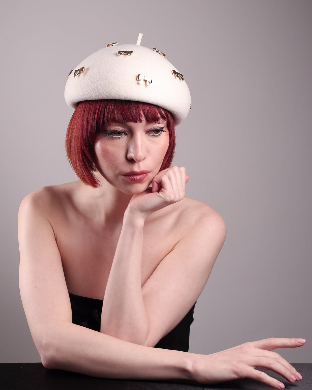 The model showcases the front view of the dome-shaped, round Elodie pillbox beret hat, crafted from soft white felt and adorned with gold coloured metal bows. Designed by Merve Bayindir.