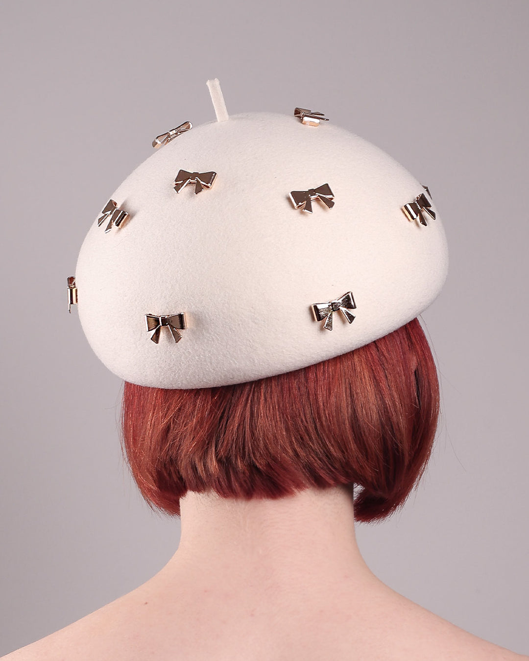 The model showcases the back close up of the dome-shaped, round Elodie pillbox beret hat, crafted from soft white felt and adorned with gold coloured metal bows. Designed by Merve Bayindir.