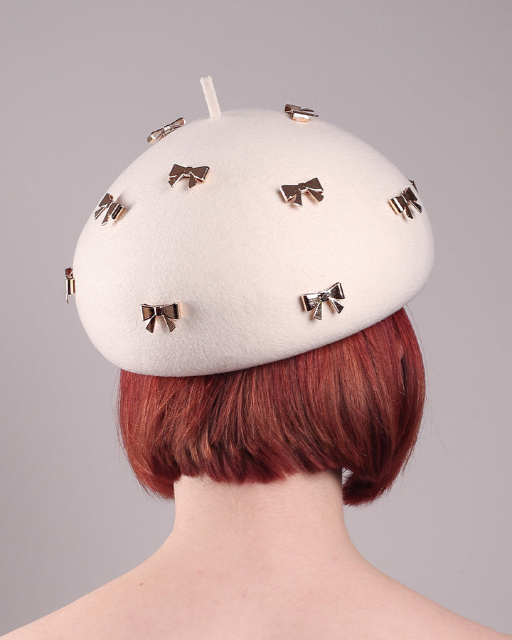 The model showcases the back close up of the dome-shaped, round Elodie pillbox beret hat, crafted from soft white felt and adorned with gold coloured metal bows. Designed by Merve Bayindir.