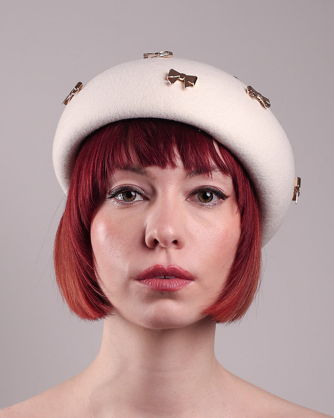 The model poses to showcase the left front angle dome-shaped, round Elodie pillbox beret hat, crafted from soft white felt and adorned with gold coloured metal bows. Designed by Merve Bayindir.