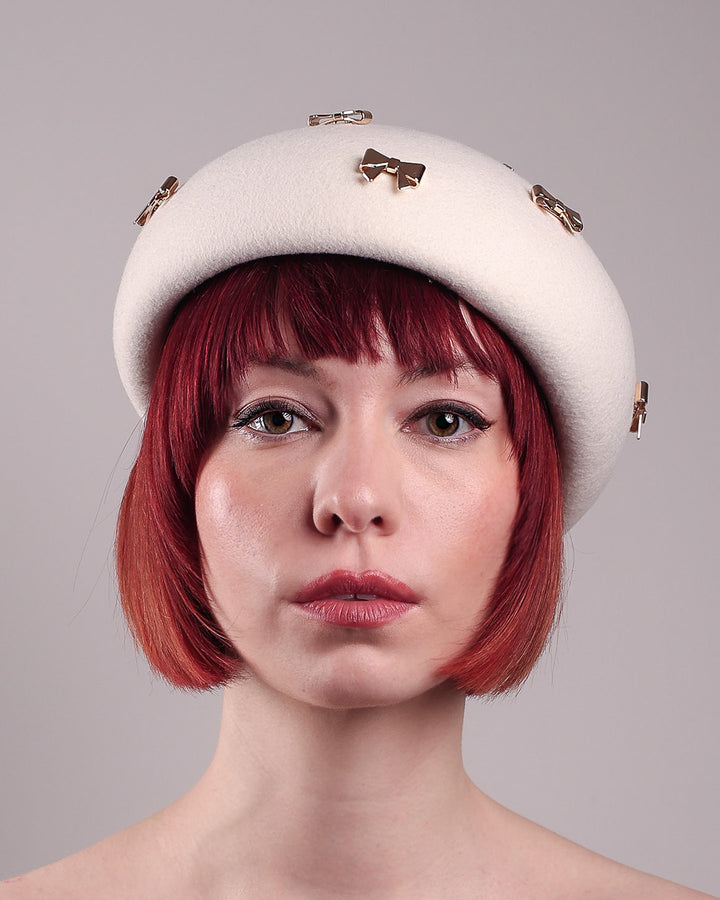 The model poses to showcase the left front angle dome-shaped, round Elodie pillbox beret hat, crafted from soft white felt and adorned with gold coloured metal bows. Designed by Merve Bayindir.