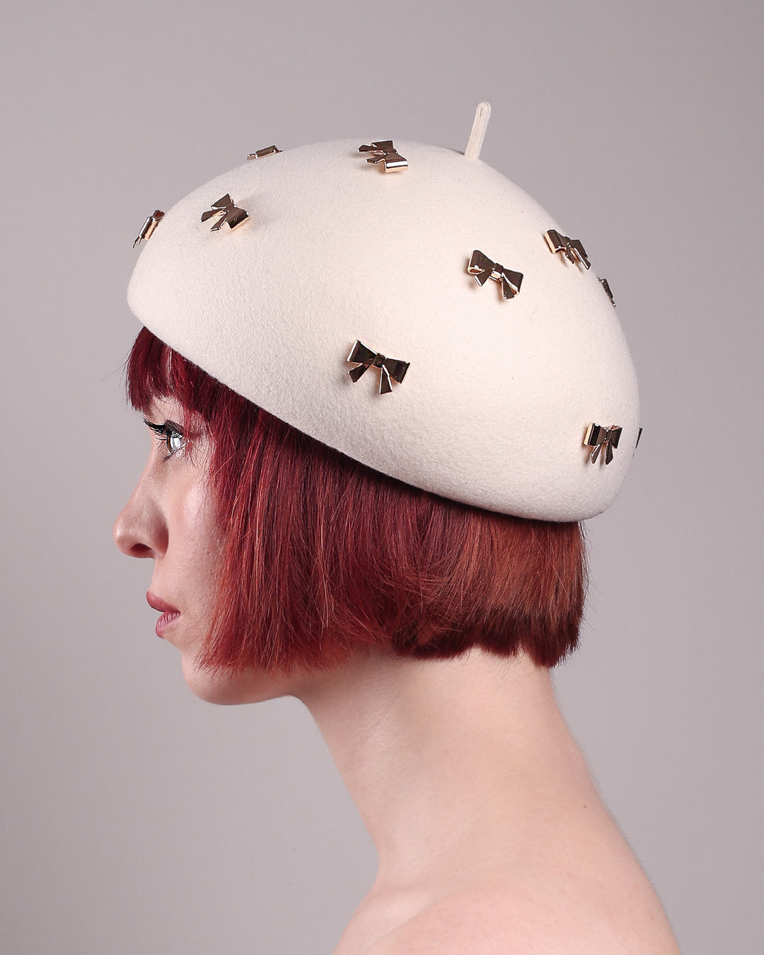 The model poses to showcase the left front angle dome-shaped, round Elodie pillbox beret hat, crafted from soft white felt and adorned with gold coloured metal bows. Designed by Merve Bayindir.