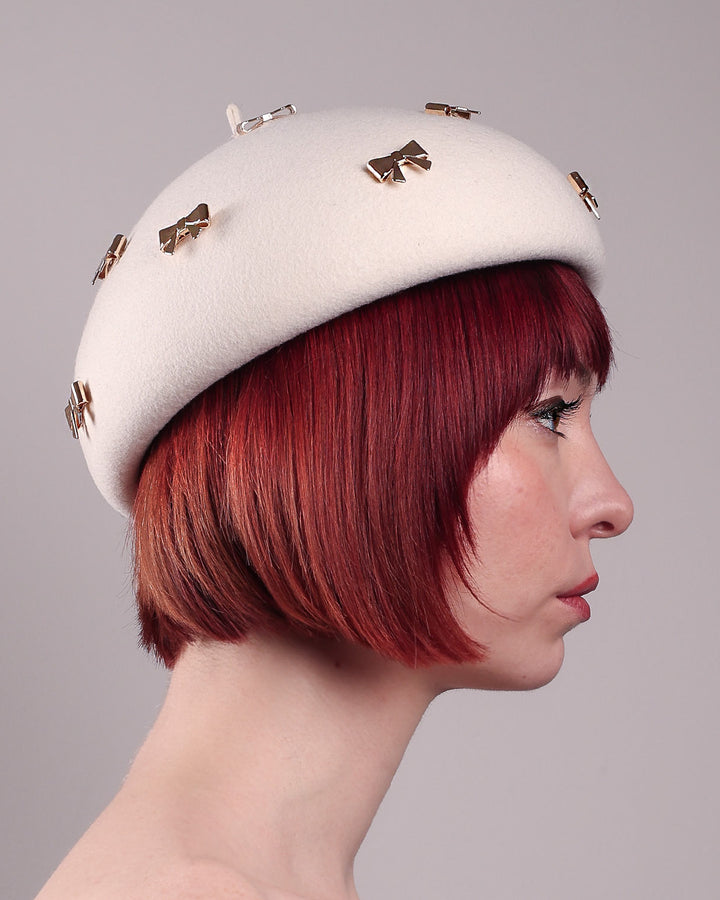 The model showcases the right side close up of the dome-shaped, round Elodie pillbox beret hat, crafted from soft white felt and adorned with gold coloured metal bows. Designed by Merve Bayindir.