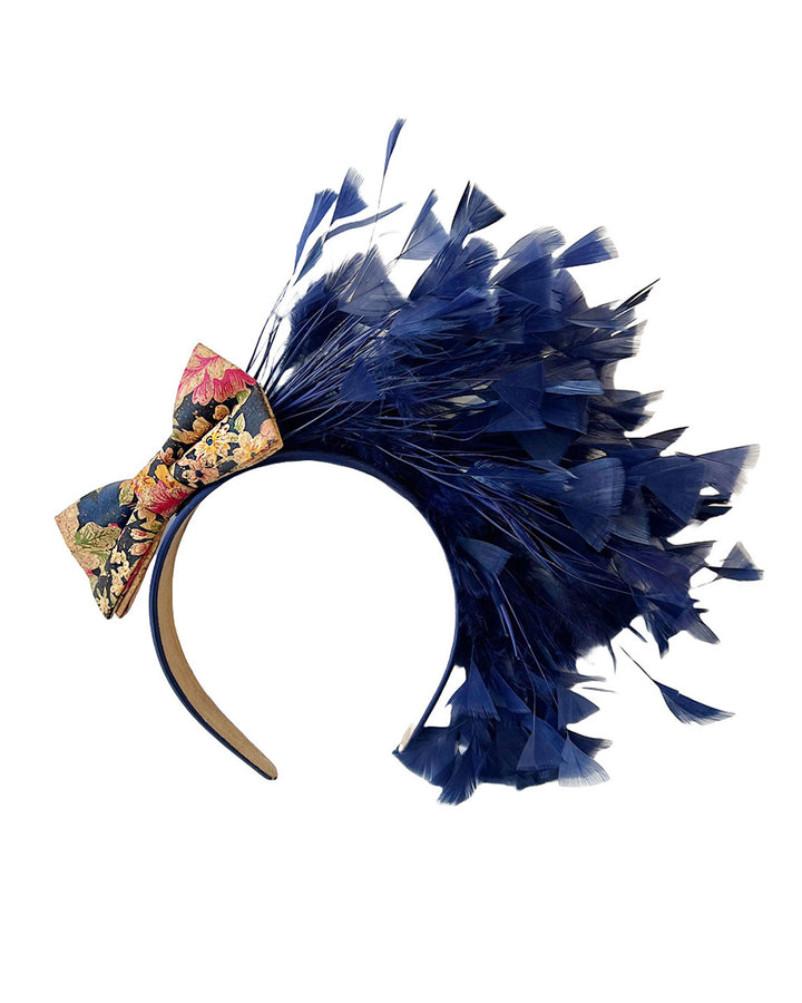 Still life picture of Esmee, where a navy storm of fine-cut feathers bursts from the same-coloured Alice headband, like smoke in reverse, anchored by a flower motif printed on a textured crock wood bow. Designed by Merve Bayindir.
