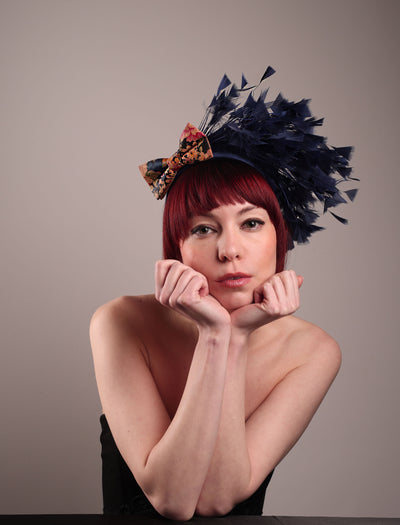 The model showcases the front of Esmee, where a navy storm of fine-cut feathers bursts from the same-coloured Alice headband, like smoke in reverse, anchored by a flower motif printed on a textured crock wood bow. Designed by Merve Bayindir.