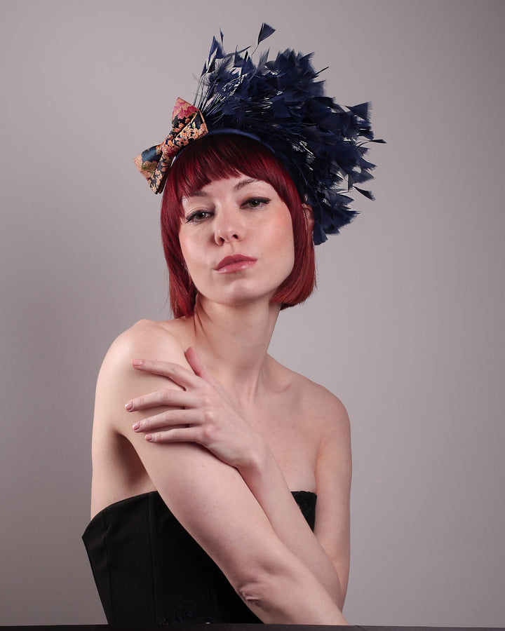 The model showcases the front left angle of Esmee, where a navy storm of fine-cut feathers bursts from the same-coloured Alice headband, like smoke in reverse, anchored by a flower motif printed on a textured crock wood bow. Designed by Merve Bayindir.