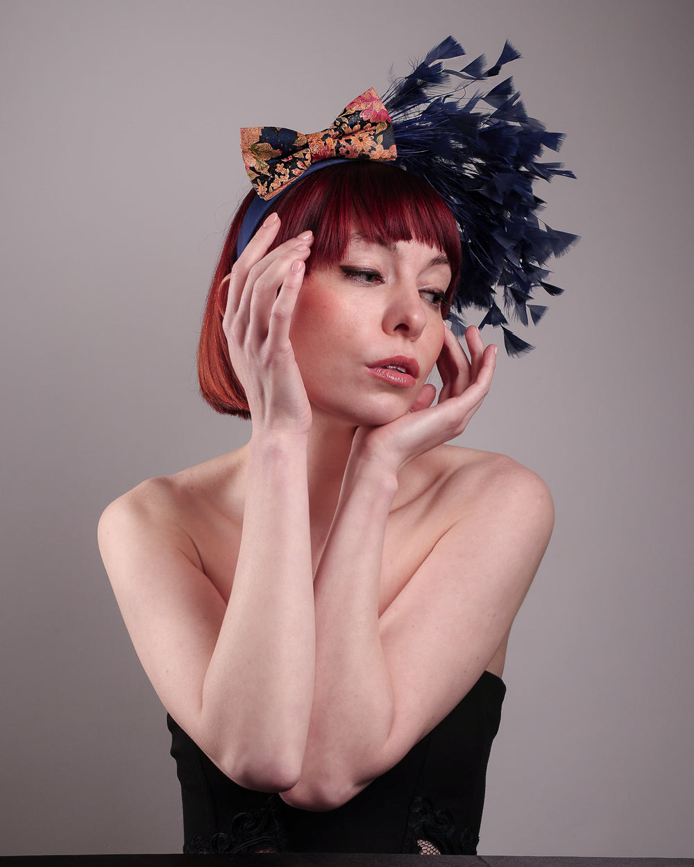 The model showcases the right side below angle of Esmee, where a navy storm of fine-cut feathers bursts from the same-coloured Alice headband, like smoke in reverse, anchored by a flower motif printed on a textured crock wood bow. Designed by Merve Bayindir.
