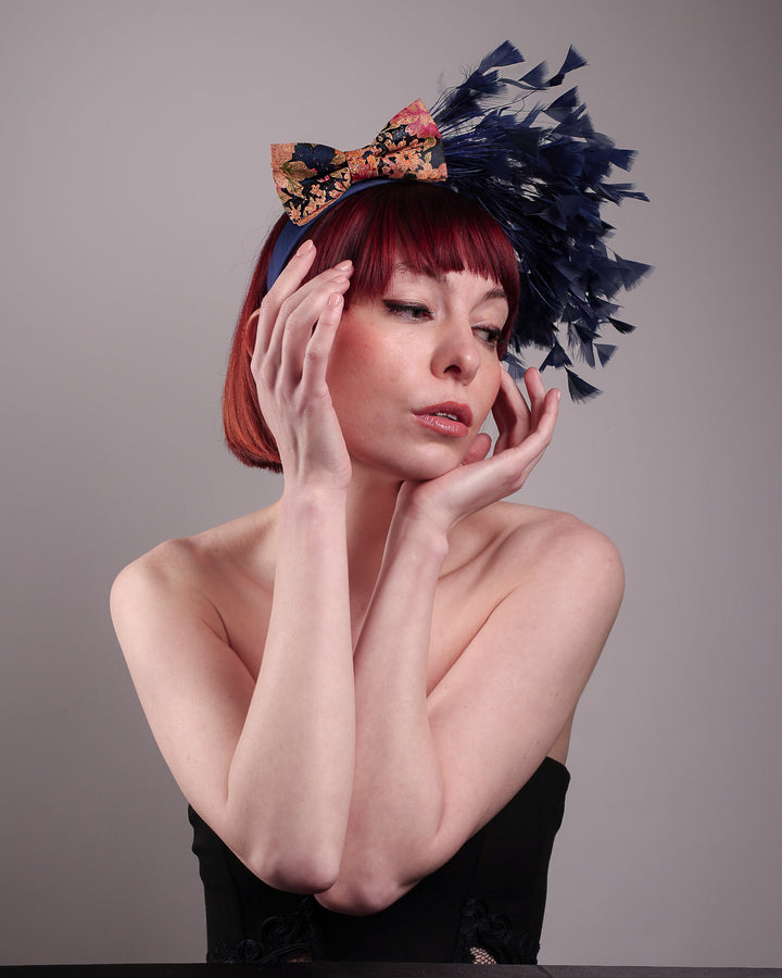 The model showcases the right side below angle of Esmee, where a navy storm of fine-cut feathers bursts from the same-coloured Alice headband, like smoke in reverse, anchored by a flower motif printed on a textured crock wood bow. Designed by Merve Bayindir.