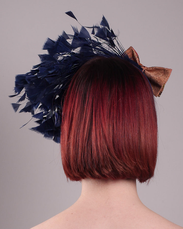 The model showcases the back of Esmee, where a navy storm of fine-cut feathers bursts from the same-coloured Alice headband, like smoke in reverse, anchored by a flower motif printed on a textured crock wood bow. Designed by Merve Bayindir.