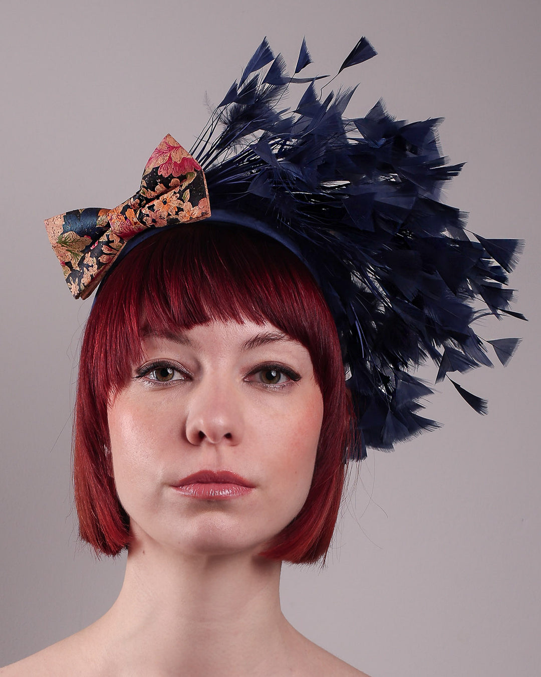 The model showcases the front close upof Esmee, where a navy storm of fine-cut feathers bursts from the same-coloured Alice headband, like smoke in reverse, anchored by a flower motif printed on a textured crock wood bow. Designed by Merve Bayindir.