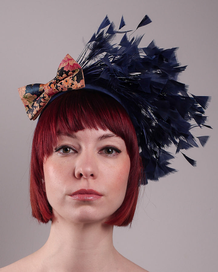 The model showcases the front close upof Esmee, where a navy storm of fine-cut feathers bursts from the same-coloured Alice headband, like smoke in reverse, anchored by a flower motif printed on a textured crock wood bow. Designed by Merve Bayindir.