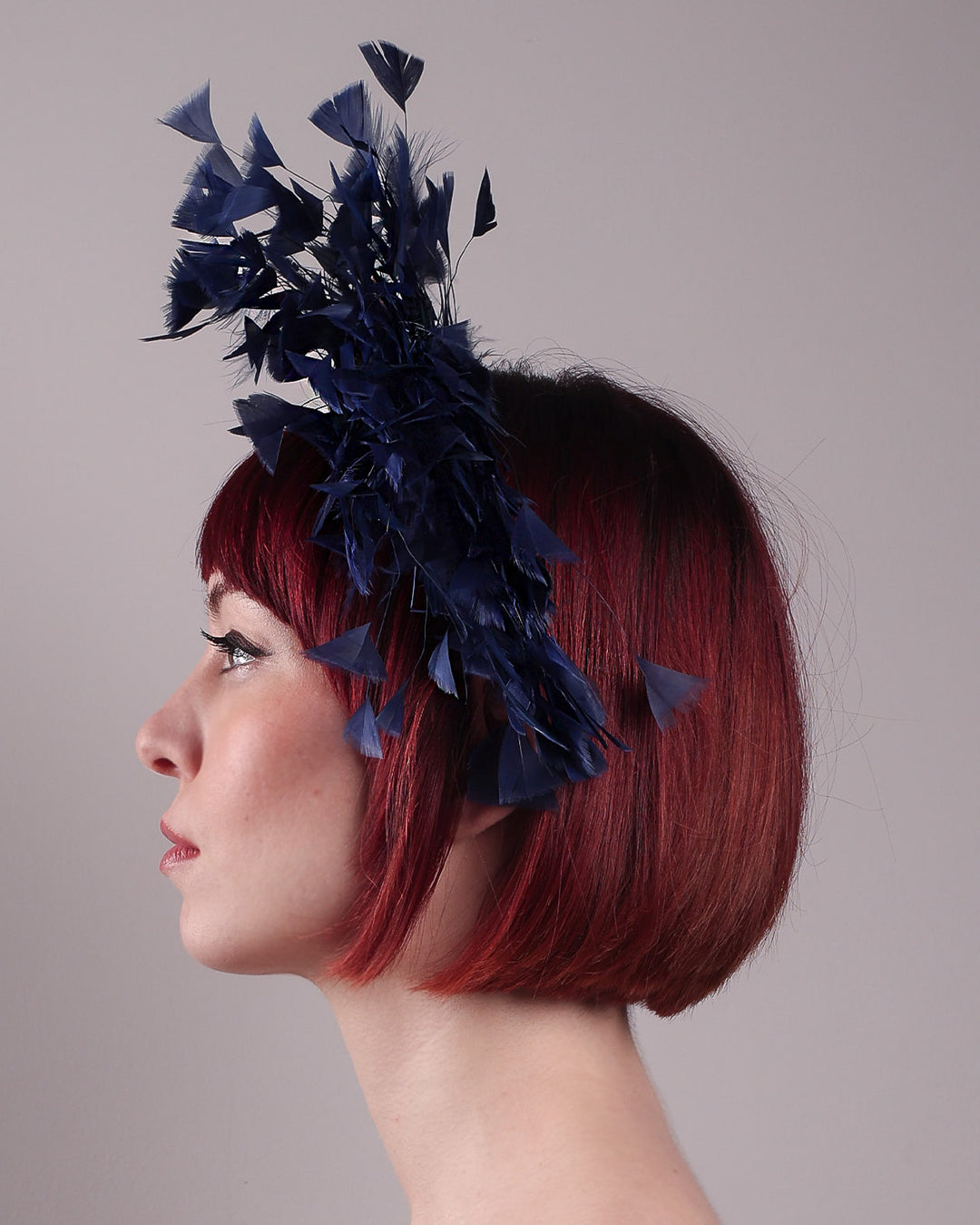 The model showcases the left side of Esmee, where a navy storm of fine-cut feathers bursts from the same-coloured Alice headband, like smoke in reverse, anchored by a flower motif printed on a textured crock wood bow. Designed by Merve Bayindir.