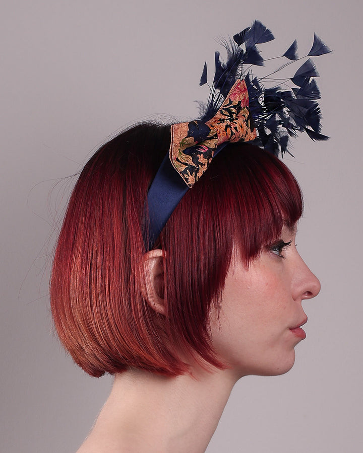 The model showcases the right side of Esmee, where a navy storm of fine-cut feathers bursts from the same-coloured Alice headband, like smoke in reverse, anchored by a flower motif printed on a textured crock wood bow. Designed by Merve Bayindir.