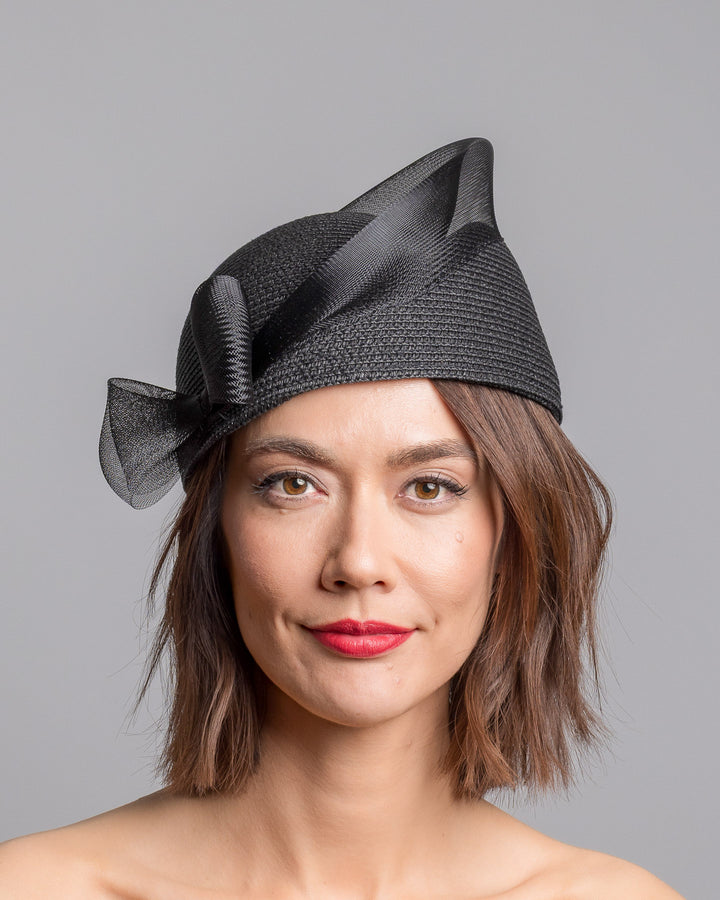 The model is showcasing the front side close up of the black straw pillbox hat with a structured woven crown, wrapped in a sheer sinamay band that curves around the top, finished with a delicate mesh bow detail at the side.Designed and made by Merve Bayindir Millinery.
