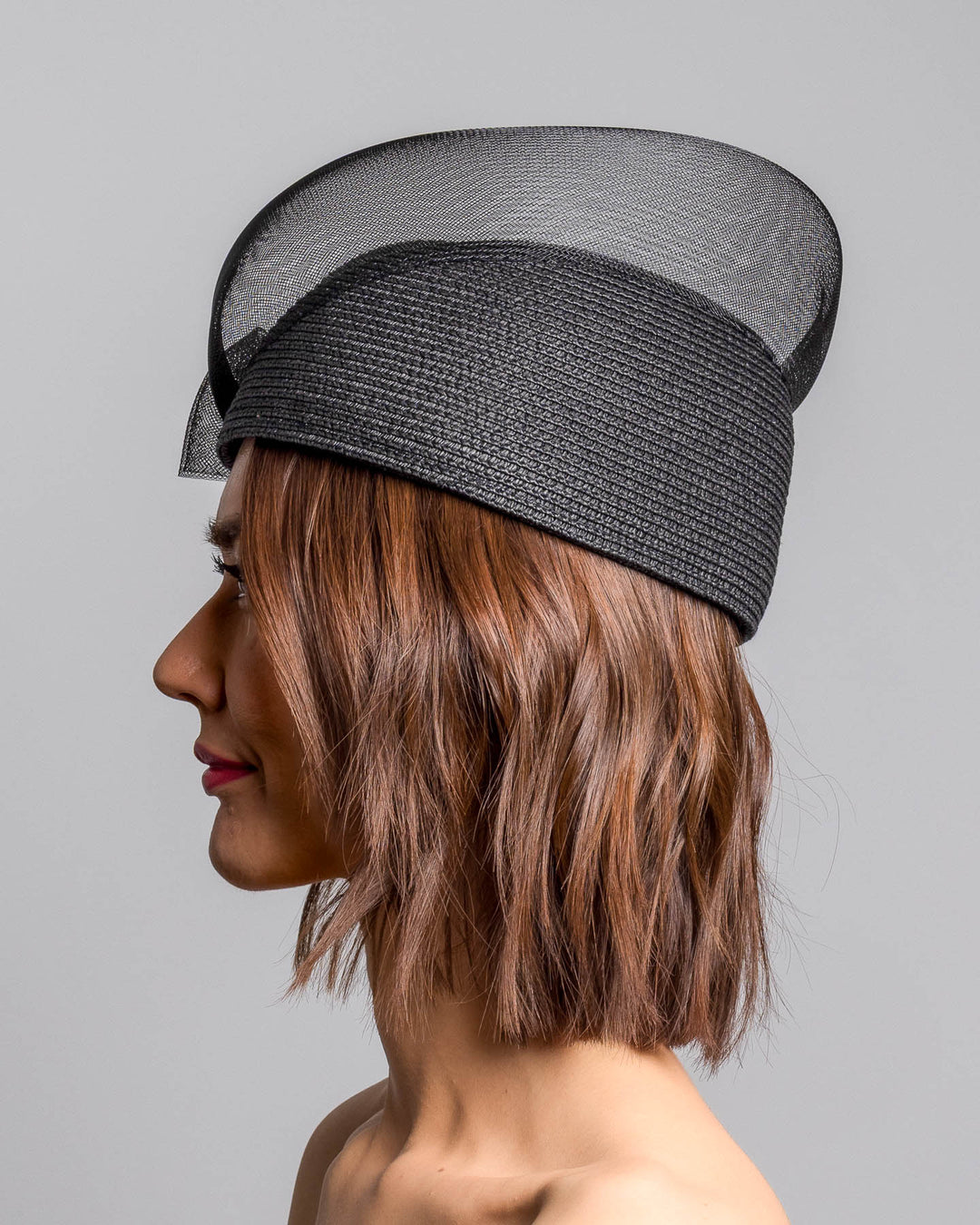 The model is showcasing the left side close up of the black straw pillbox hat with a structured woven crown, wrapped in a sheer sinamay band that curves around the top, finished with a delicate mesh bow detail at the side.Designed and made by Merve Bayindir Millinery.
