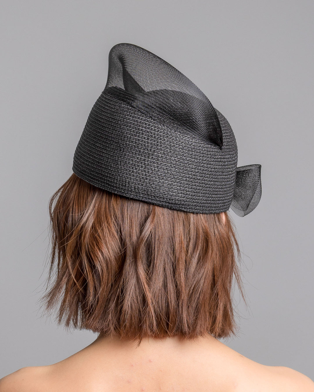 The model is showcasing the back side close up of the black straw pillbox hat with a structured woven crown, wrapped in a sheer sinamay band that curves around the top, finished with a delicate mesh bow detail at the side.Designed and made by Merve Bayindir Millinery.
