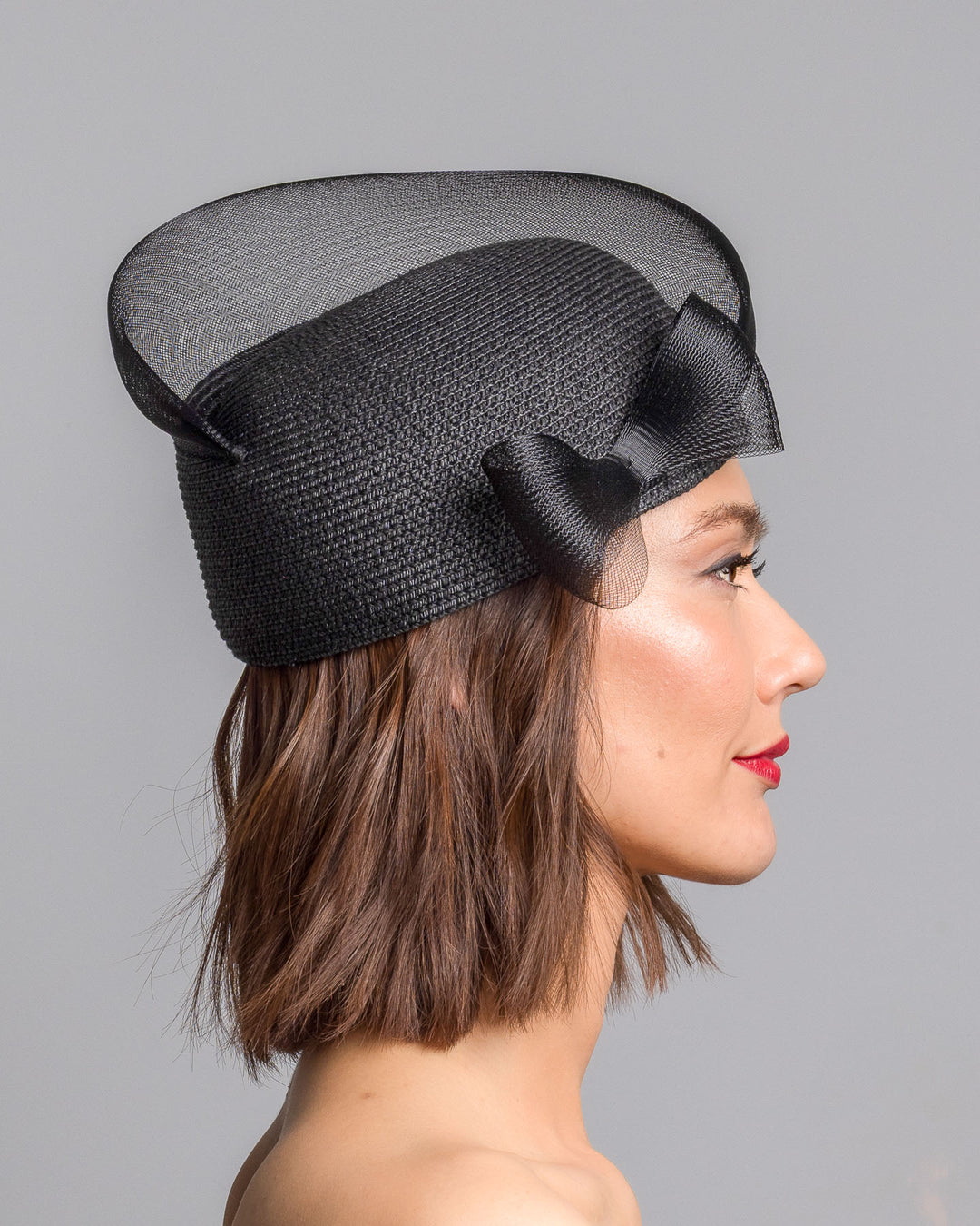 The model is showcasing the right side close up of the black straw pillbox hat with a structured woven crown, wrapped in a sheer sinamay band that curves around the top, finished with a delicate mesh bow detail at the side.Designed and made by Merve Bayindir Millinery.
