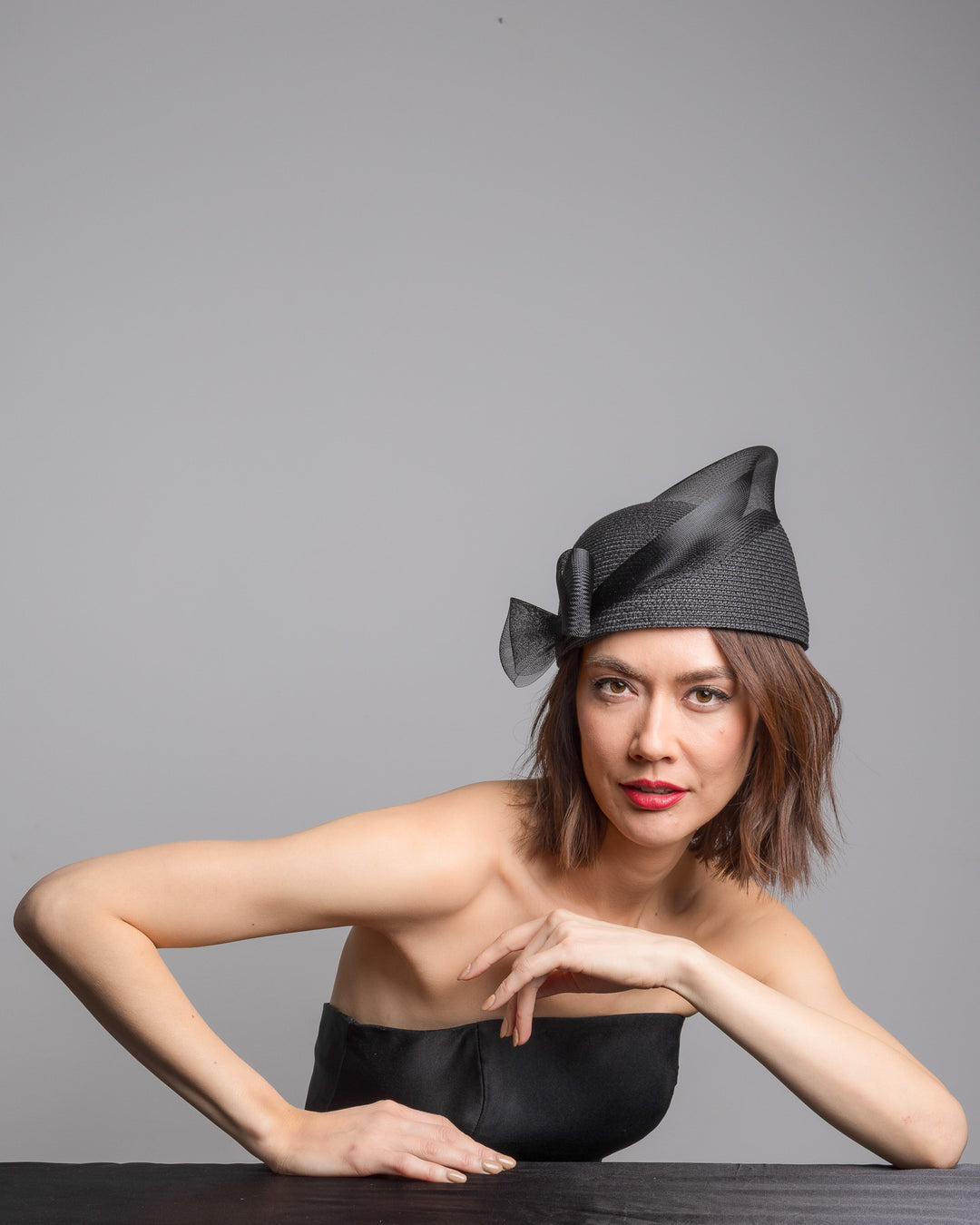 The female model is posing, wearing 
Fayne, the black straw pillbox hat with a structured woven crown, wrapped in a sheer sinamay band that curves around the top, finished with a delicate mesh bow detail at the side.Designed and made by Merve Bayindir Millinery.
