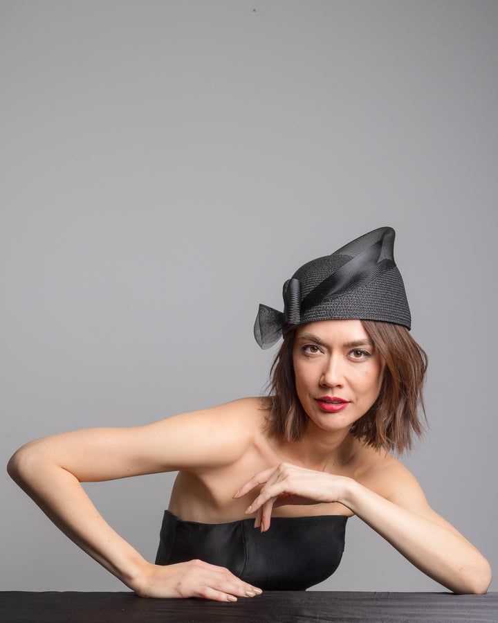 The female model is posing, wearing 
Fayne, the black straw pillbox hat with a structured woven crown, wrapped in a sheer sinamay band that curves around the top, finished with a delicate mesh bow detail at the side.Designed and made by Merve Bayindir Millinery.
