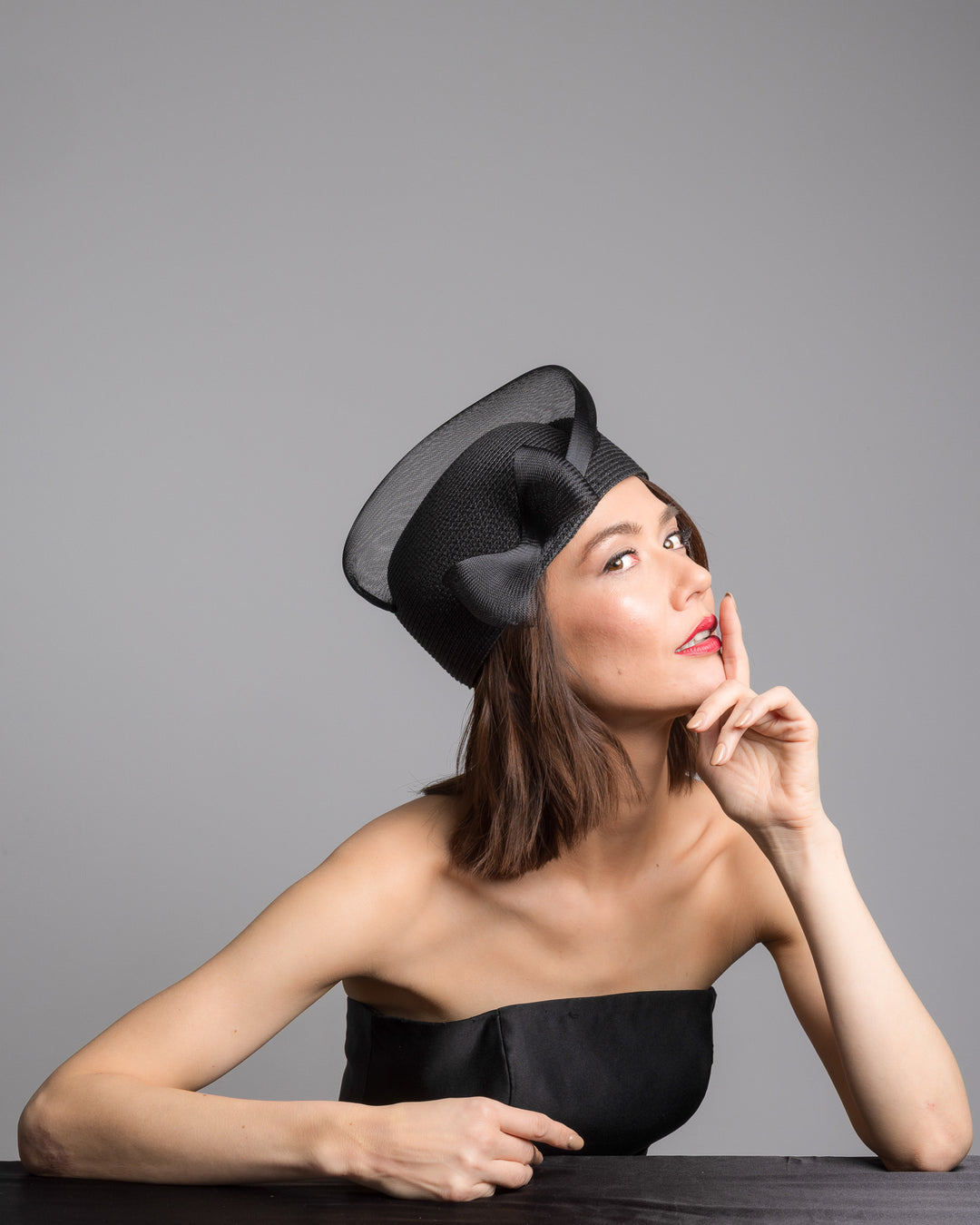 The female model is posing, wearing 
Fayne, the black straw pillbox hat with a structured woven crown, wrapped in a sheer sinamay band that curves around the top, finished with a delicate mesh bow detail at the side.Designed and made by Merve Bayindir Millinery.
