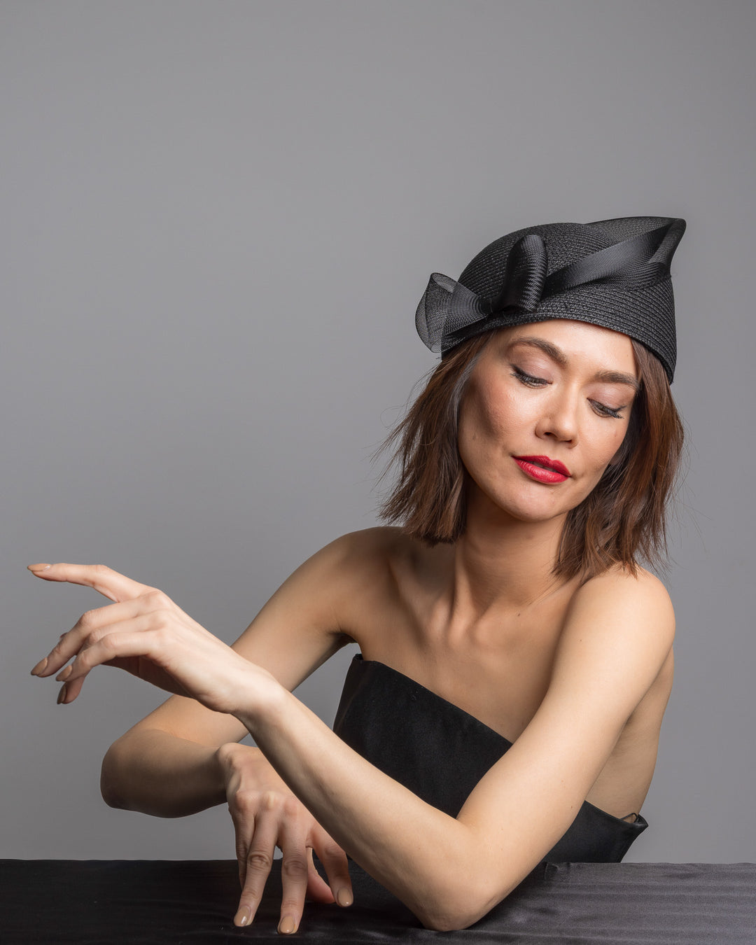 The female model is posing, wearing 
Fayne, the black straw pillbox hat with a structured woven crown, wrapped in a sheer sinamay band that curves around the top, finished with a delicate mesh bow detail at the side.Designed and made by Merve Bayindir Millinery.
