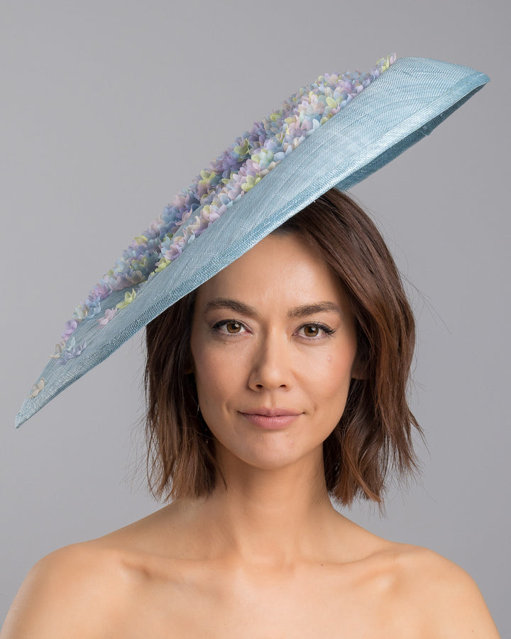 The model is showcasing the front side close up of the powder blue sinamay base hat with a wide, gently sloping brim, adorned with clusters of soft pastel organza flowers in lilac, pale yellow, and blush tones that gather along the crown and trail lightly across the surface. Designed and made by Merve Bayindir Millinery.