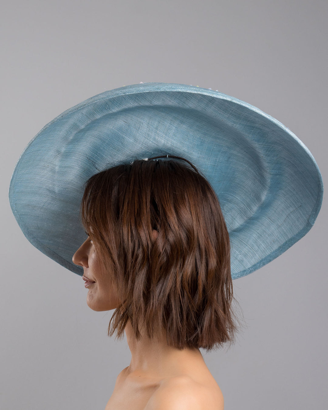 The model is showcasing the left side close up of the powder blue sinamay base hat with a wide, gently sloping brim, adorned with clusters of soft pastel organza flowers in lilac, pale yellow, and blush tones that gather along the crown and trail lightly across the surface. Designed and made by Merve Bayindir Millinery.