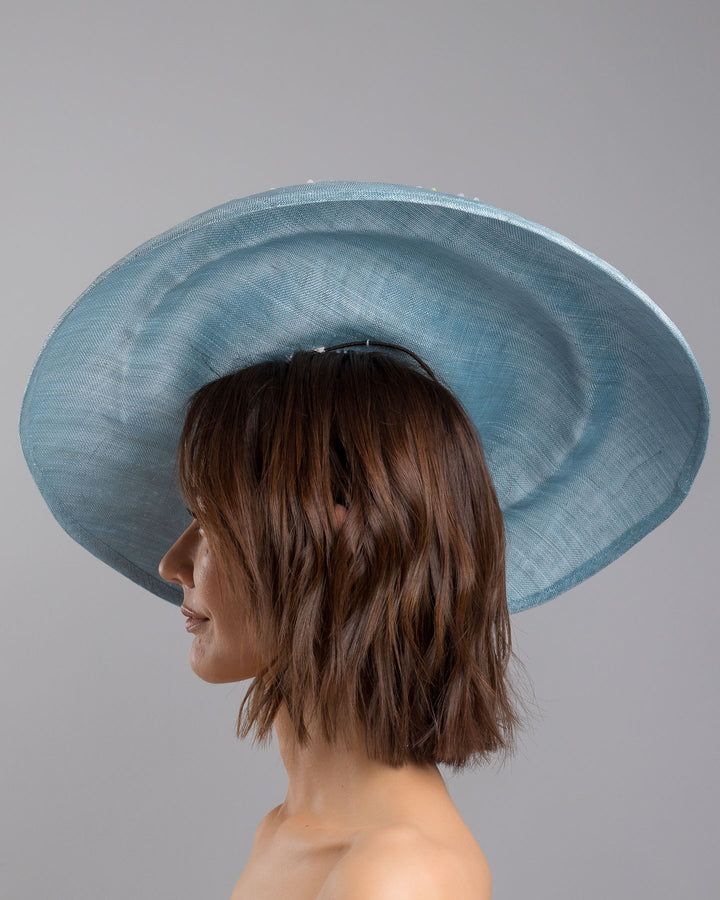 The model is showcasing the left side close up of the powder blue sinamay base hat with a wide, gently sloping brim, adorned with clusters of soft pastel organza flowers in lilac, pale yellow, and blush tones that gather along the crown and trail lightly across the surface. Designed and made by Merve Bayindir Millinery.