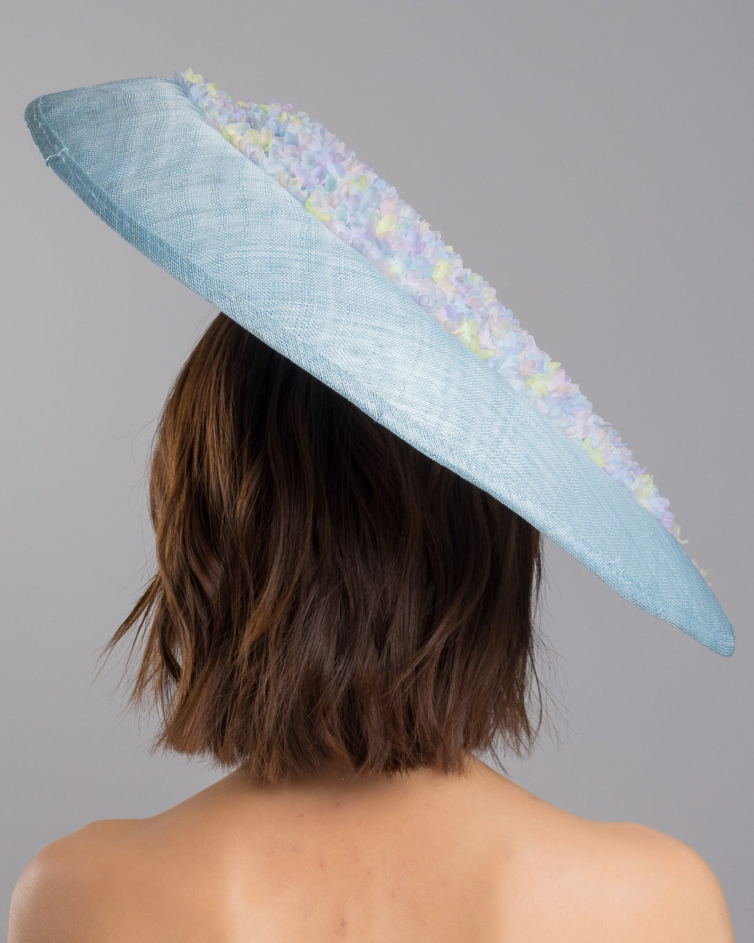 The model is showcasing the back side close up of the powder blue sinamay base hat with a wide, gently sloping brim, adorned with clusters of soft pastel organza flowers in lilac, pale yellow, and blush tones that gather along the crown and trail lightly across the surface. Designed and made by Merve Bayindir Millinery.