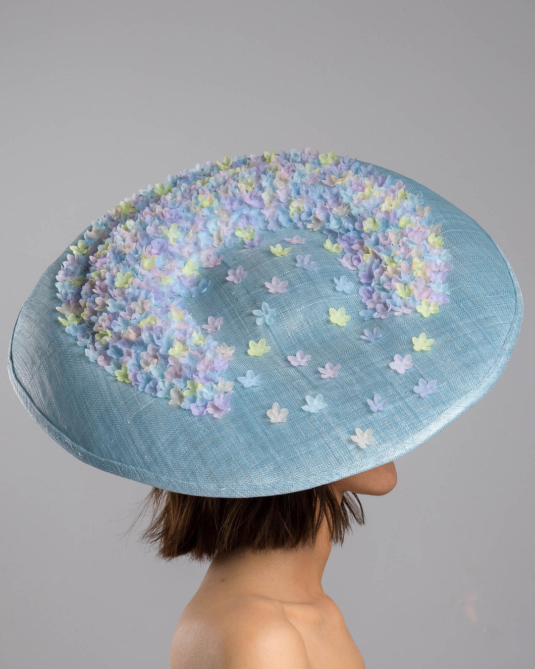 The model is showcasing the right side close up of the powder blue sinamay base hat with a wide, gently sloping brim, adorned with clusters of soft pastel organza flowers in lilac, pale yellow, and blush tones that gather along the crown and trail lightly across the surface. Designed and made by Merve Bayindir Millinery.
