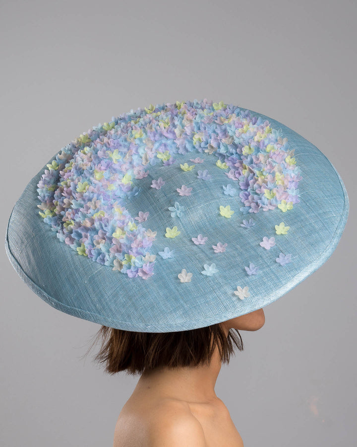 The model is showcasing the right side close up of the powder blue sinamay base hat with a wide, gently sloping brim, adorned with clusters of soft pastel organza flowers in lilac, pale yellow, and blush tones that gather along the crown and trail lightly across the surface. Designed and made by Merve Bayindir Millinery.
