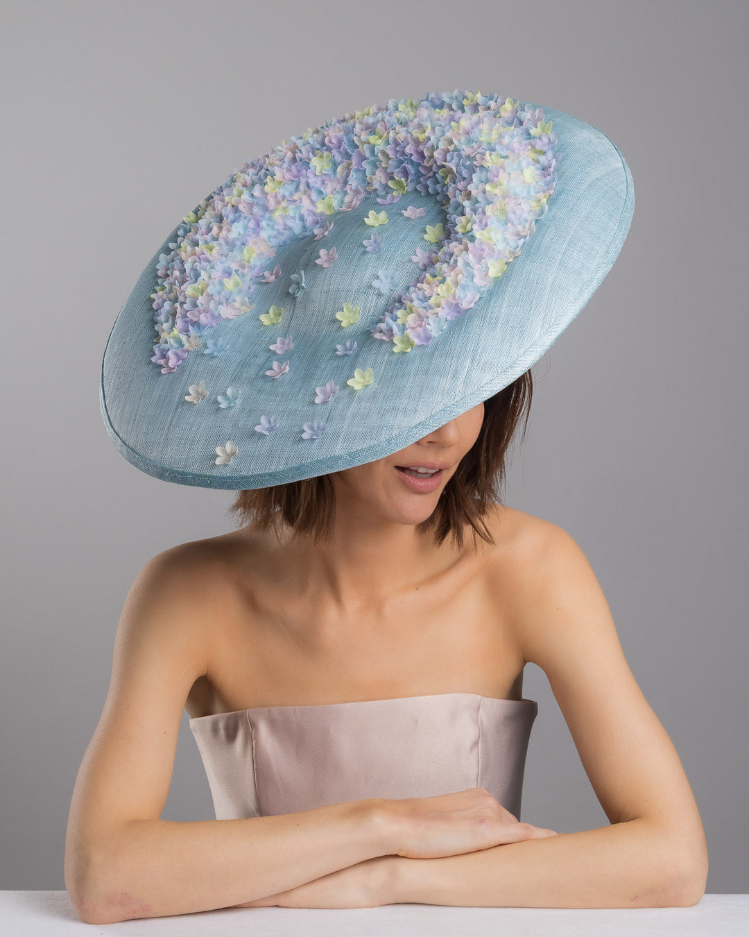 The female model is posing, wearing
Feyra, the powder blue sinamay base hat with a wide, gently sloping brim, adorned with clusters of soft pastel organza flowers in lilac, pale yellow, and blush tones that gather along the crown and trail lightly across the surface. Designed and made by Merve Bayindir Millinery.