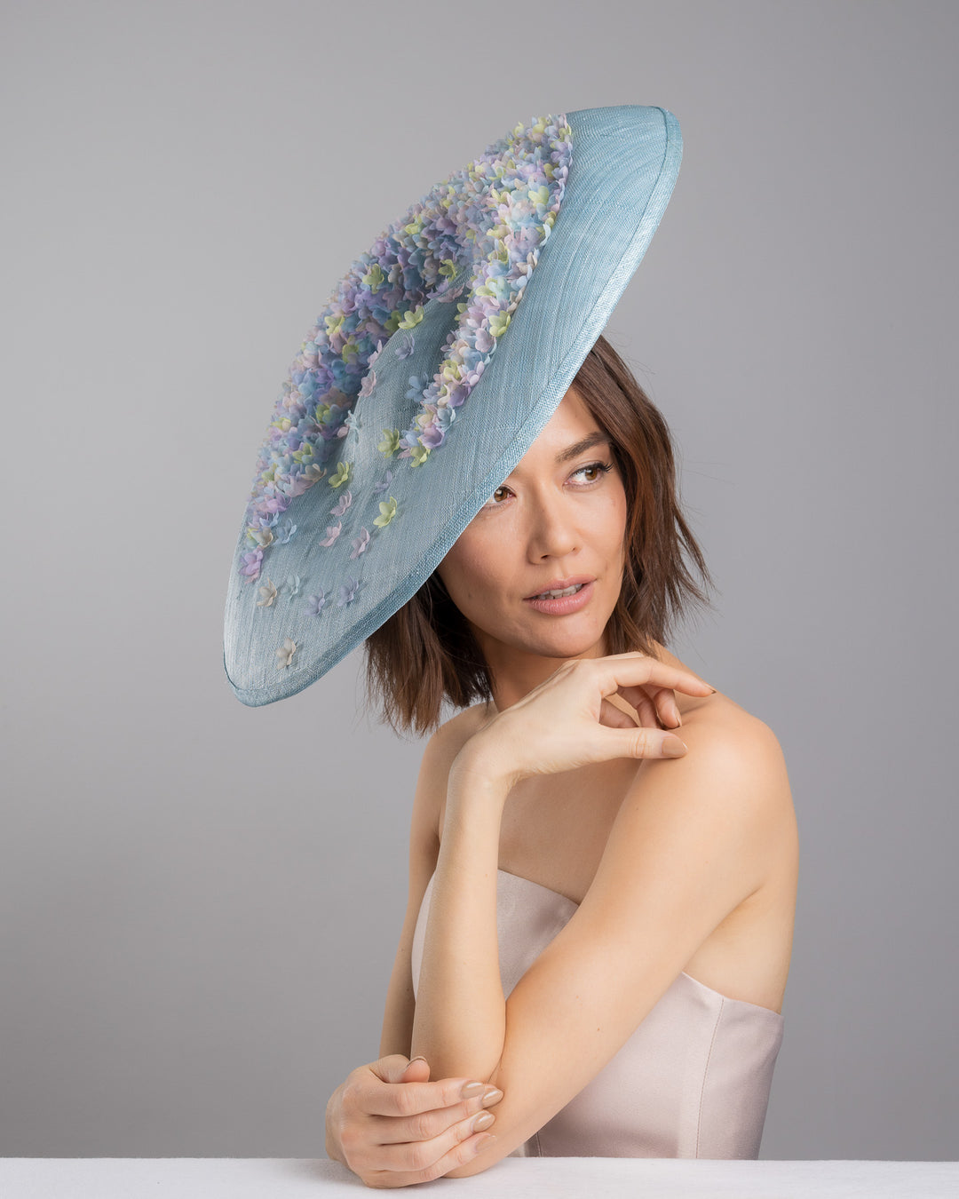 The female model is posing, wearing
Feyra, the powder blue sinamay base hat with a wide, gently sloping brim, adorned with clusters of soft pastel organza flowers in lilac, pale yellow, and blush tones that gather along the crown and trail lightly across the surface. Designed and made by Merve Bayindir Millinery.