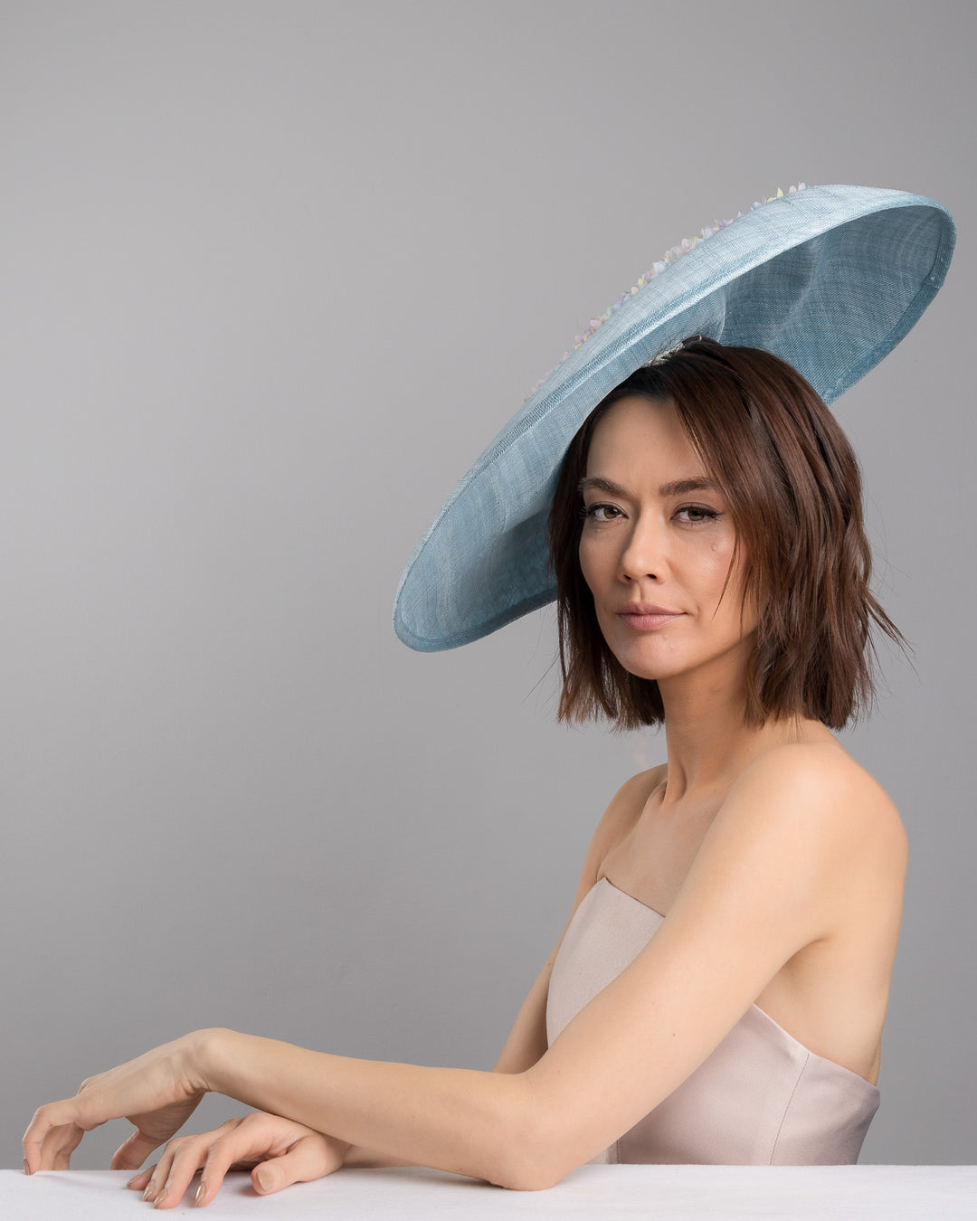 The female model is posing, wearing
Feyra, the powder blue sinamay base hat with a wide, gently sloping brim, adorned with clusters of soft pastel organza flowers in lilac, pale yellow, and blush tones that gather along the crown and trail lightly across the surface. Designed and made by Merve Bayindir Millinery.