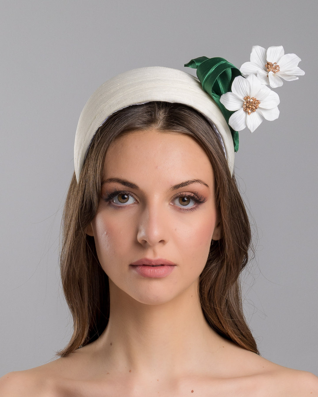 The model is showcasing the front side close up of the sructured white half-hat in smooth fabric, adorned with two white floral appliqués featuring gold beaded centers and finished with sculpted deep green leaves for contrast. Designed and made by Merve Bayindir Millinery.
