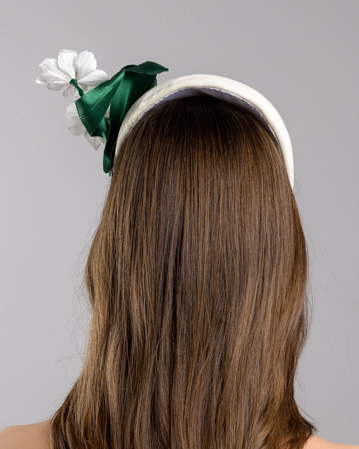 The model is showcasing the back side close up of the sructured white half-hat in smooth fabric, adorned with two white floral appliqués featuring gold beaded centers and finished with sculpted deep green leaves for contrast. Designed and made by Merve Bayindir Millinery.
