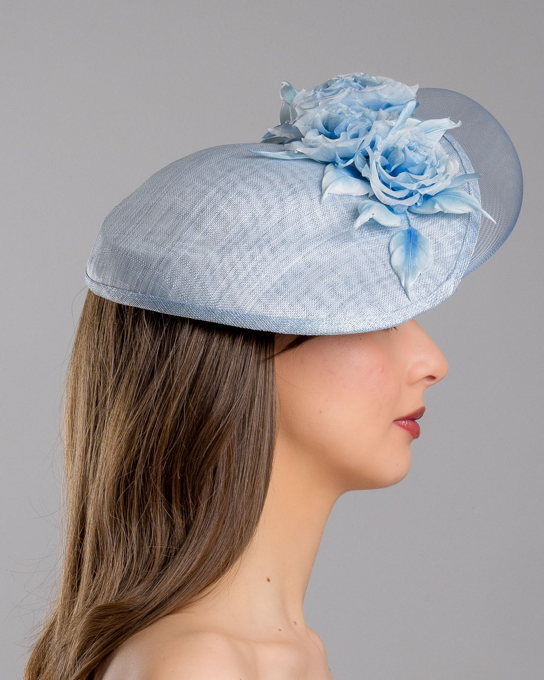 The model is showcasing the right side close up of the powder blue sinamay fascinator with a sculpted teardrop silhouette, adorned with layered chiffon roses and matching soft leaves for an elegant, romantic finish. Designed and made by Merve Bayindir Millinery.
