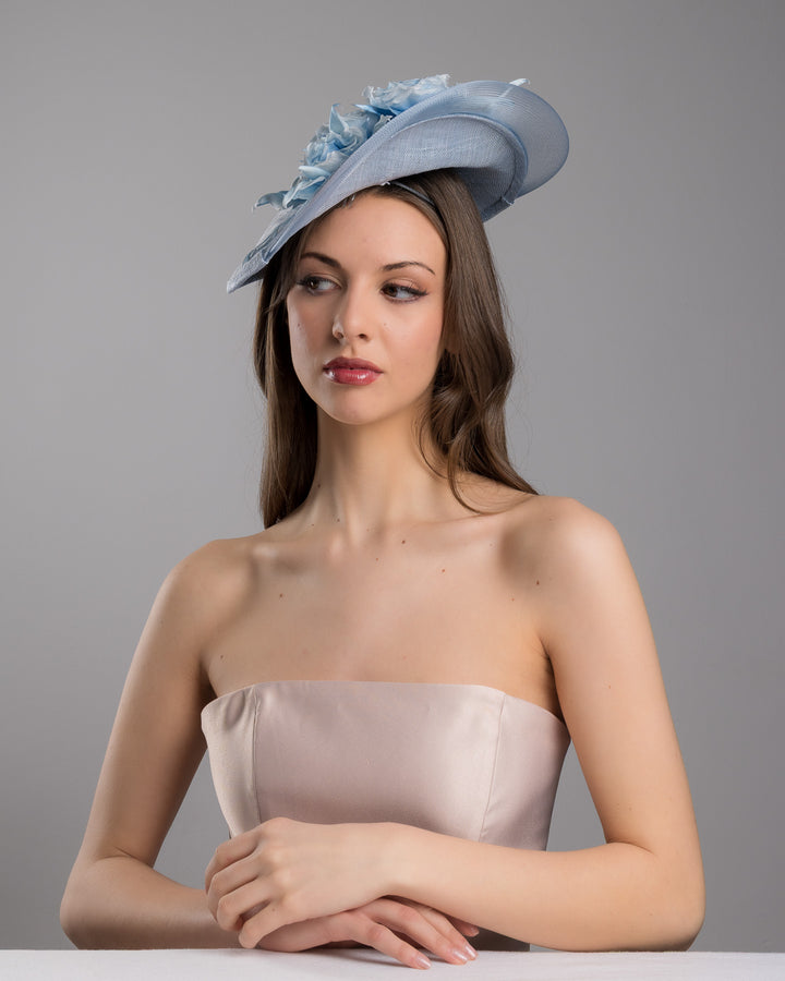 The female model is posing, wearing
Frey,  the powder blue sinamay fascinator with a sculpted teardrop silhouette, adorned with layered chiffon roses and matching soft leaves for an elegant, romantic finish. Designed and made by Merve Bayindir Millinery.
