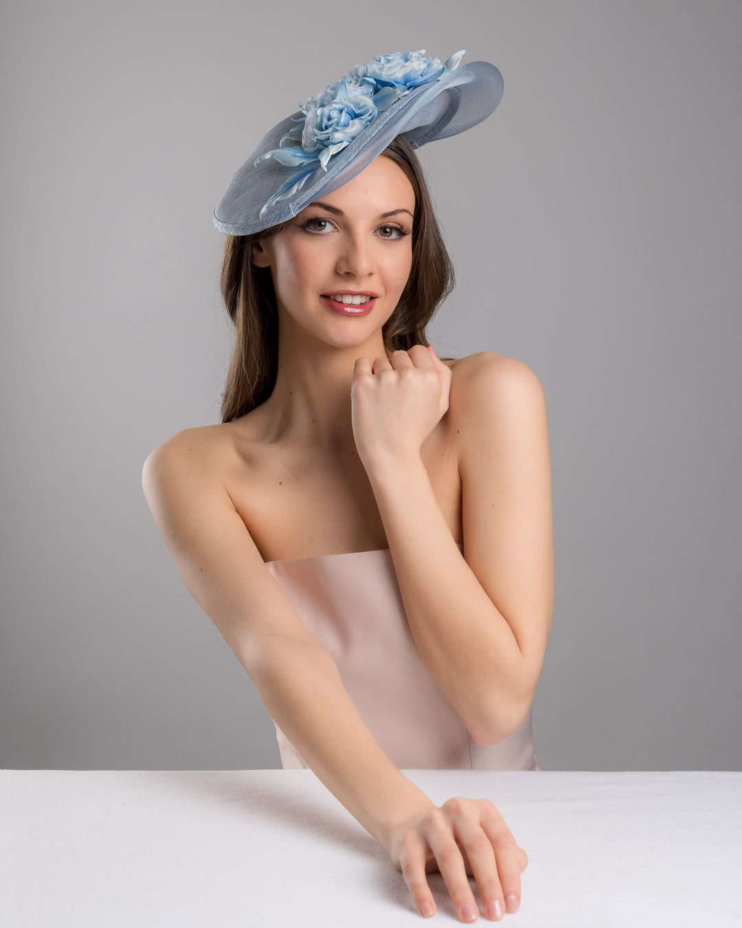 The female model is posing, wearing
Frey,  the powder blue sinamay fascinator with a sculpted teardrop silhouette, adorned with layered chiffon roses and matching soft leaves for an elegant, romantic finish. Designed and made by Merve Bayindir Millinery.
