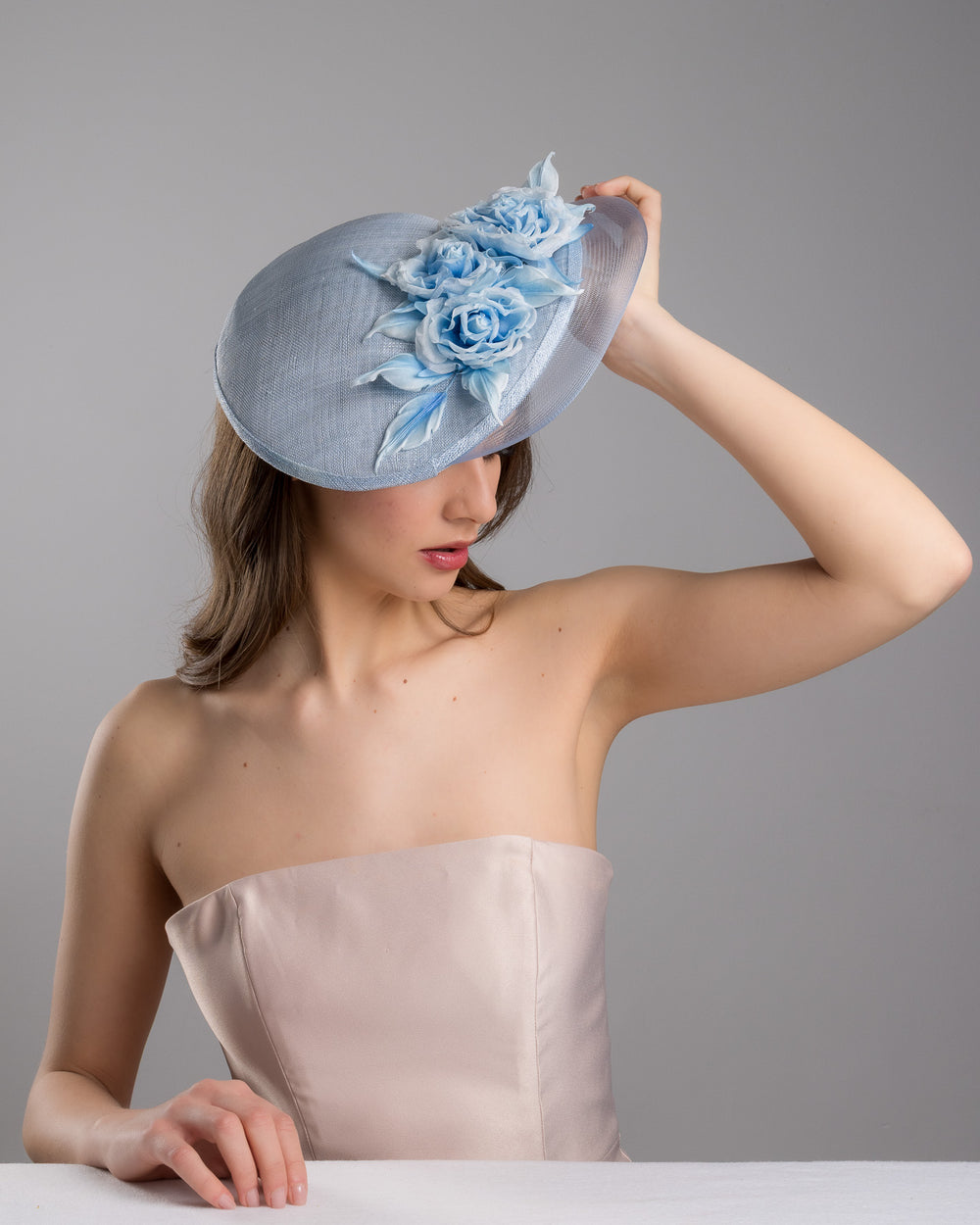 The female model is posing, wearing
Frey,  the powder blue sinamay fascinator with a sculpted teardrop silhouette, adorned with layered chiffon roses and matching soft leaves for an elegant, romantic finish. Designed and made by Merve Bayindir Millinery.
